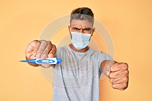 Young hispanic man wearing medical mask holding thermometer annoyed and frustrated shouting with anger, yelling crazy with anger