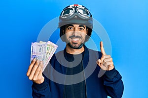 Young hispanic man wearing motorcycle helmet holding indian rupee smiling with an idea or question pointing finger with happy
