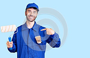 Young hispanic man wearing painter uniform holding roller pointing finger to one self smiling happy and proud