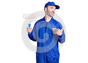 Young hispanic man wearing painter uniform holding roller pointing thumb up to the side smiling happy with open mouth