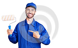 Young hispanic man wearing painter uniform holding roller smiling happy pointing with hand and finger