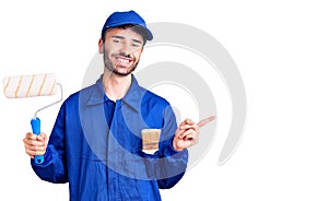 Young hispanic man wearing painter uniform holding roller smiling happy pointing with hand and finger to the side