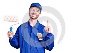 Young hispanic man wearing painter uniform holding roller surprised with an idea or question pointing finger with happy face,