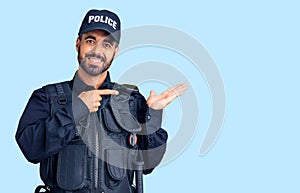 Young hispanic man wearing police uniform amazed and smiling to the camera while presenting with hand and pointing with finger