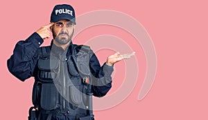 Young hispanic man wearing police uniform confused and annoyed with open palm showing copy space and pointing finger to forehead