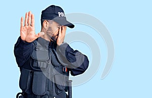 Young hispanic man wearing police uniform covering eyes with hands and doing stop gesture with sad and fear expression