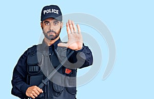 Young hispanic man wearing police uniform holding baton with open hand doing stop sign with serious and confident expression,