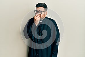Young hispanic man wearing priest uniform standing over white background smelling something stinky and disgusting, intolerable