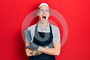 Young hispanic man wearing professional apron holding knife angry and mad screaming frustrated and furious, shouting with anger