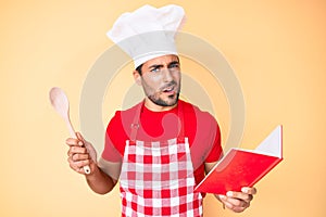 Young hispanic man wearing professional baker apron reading cooking recipe book clueless and confused expression