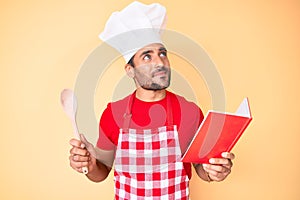 Young hispanic man wearing professional baker apron reading cooking recipe book smiling looking to the side and staring away
