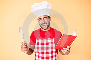 Young hispanic man wearing professional baker apron reading cooking recipe book winking looking at the camera with sexy