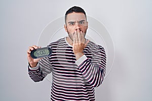 Young hispanic man wearing pyjama holding alarm clock covering mouth with hand, shocked and afraid for mistake