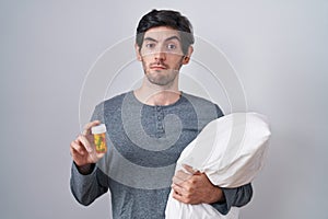 Young hispanic man wearing pyjama hugging pillow holding pills clueless and confused expression