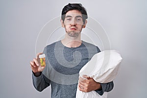 Young hispanic man wearing pyjama hugging pillow holding pills puffing cheeks with funny face