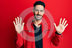 Young hispanic man wearing red leather jacket showing and pointing up with fingers number ten while smiling confident and happy