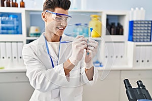 Young hispanic man wearing scientist uniform writing on test tube at laboratory