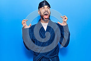 Young hispanic man wearing sleep mask and robe crazy and mad shouting and yelling with aggressive expression and arms raised