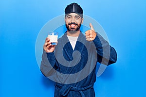 Young hispanic man wearing sleep mask and robe drinking milk smiling happy and positive, thumb up doing excellent and approval