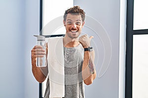 Young hispanic man wearing sportswear drinking water pointing to the back behind with hand and thumbs up, smiling confident