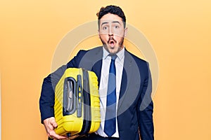 Young hispanic man wearing suit holding cabin bag scared and amazed with open mouth for surprise, disbelief face