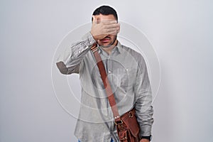 Young hispanic man wearing suitcase covering eyes with hand, looking serious and sad