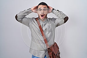 Young hispanic man wearing suitcase crazy and scared with hands on head, afraid and surprised of shock with open mouth