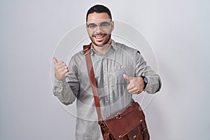 Young hispanic man wearing suitcase pointing to the back behind with hand and thumbs up, smiling confident