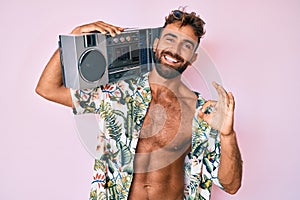Young hispanic man wearing summer clothes holding boombox, listening to music smiling in love doing heart symbol shape with hands