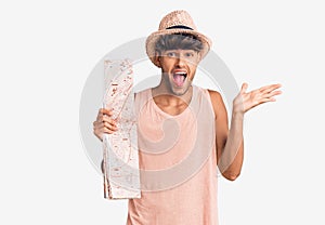 Young hispanic man wearing summer hat holding map celebrating victory with happy smile and winner expression with raised hands