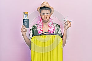 Young hispanic man wearing summer style and hawaiian lei holding passport and plane toy depressed and worry for distress, crying