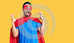 Young hispanic man wearing super hero costume crazy and mad shouting and yelling with aggressive expression and arms raised