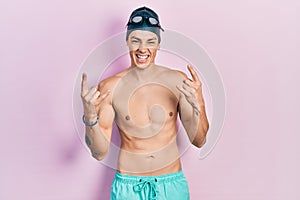 Young hispanic man wearing swimwear and swimmer glasses shouting with crazy expression doing rock symbol with hands up