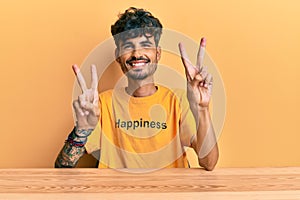 Young hispanic man wearing tshirt with happiness word message sitting on the table smiling looking to the camera showing fingers