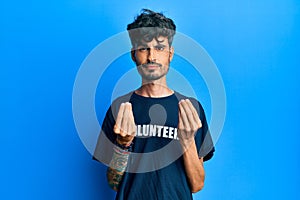 Young hispanic man wearing volunteer t shirt doing money gesture with hands, asking for salary payment, millionaire business