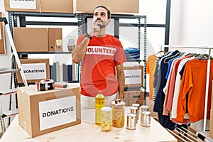 Young hispanic man wearing volunteer t shirt at donations stand looking confident at the camera smiling with crossed arms and hand