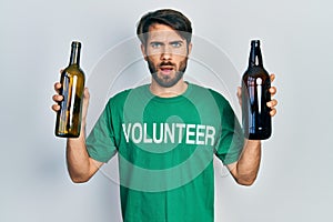 Young hispanic man wearing volunteer t shirt holding recycling bottle glass afraid and shocked with surprise and amazed