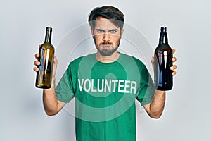 Young hispanic man wearing volunteer t shirt holding recycling bottle glass puffing cheeks with funny face