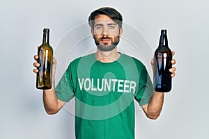 Young hispanic man wearing volunteer t shirt holding recycling bottle glass relaxed with serious expression on face
