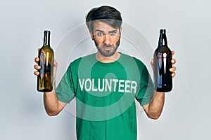 Young hispanic man wearing volunteer t shirt holding recycling bottle glass skeptic and nervous, frowning upset because of problem