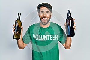 Young hispanic man wearing volunteer t shirt holding recycling bottle glass smiling and laughing hard out loud because funny crazy