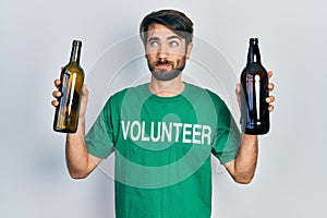 Young hispanic man wearing volunteer t shirt holding recycling bottle glass smiling looking to the side and staring away thinking