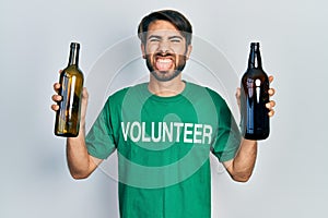 Young hispanic man wearing volunteer t shirt holding recycling bottle glass sticking tongue out happy with funny expression