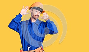 Young hispanic man wearing worker uniform doing bunny ears gesture with hands palms looking cynical and skeptical