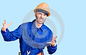 Young hispanic man wearing worker uniform shouting frustrated with rage, hands trying to strangle, yelling mad