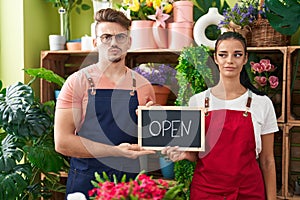 Young hispanic man a woman working at florist holding open sign smiling looking to the side and staring away thinking