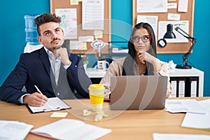 Young hispanic man and woman working at the office serious face thinking about question with hand on chin, thoughtful about