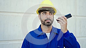 Young hispanic man worker wearing hardhat listening to voice message by smartphone at construction place