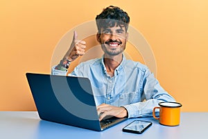 Young hispanic man working at the office drinking a cup of coffee smiling happy and positive, thumb up doing excellent and