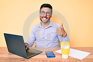 Young hispanic man working at the office drinking a cup of coffee smiling happy and positive, thumb up doing excellent and